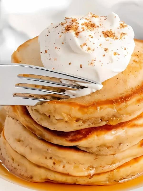 A stack of pancakes drizzled with syrup, topped with a dollop of whipped cream and a sprinkle of cinnamon. A fork is about to cut into the pancakes. The background includes a cup of coffee and cinnamon sticks.