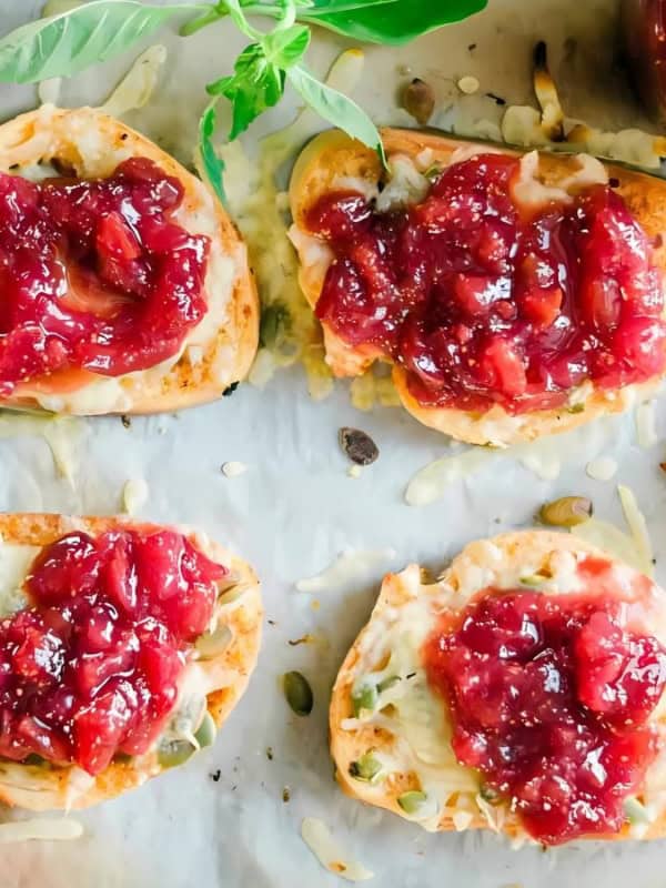 Toast slices topped with strawberry jam are arranged on parchment paper. Fresh basil leaves are scattered around, and there's a jar of jam and a spoon nearby. A daisy flower is visible on the side.
