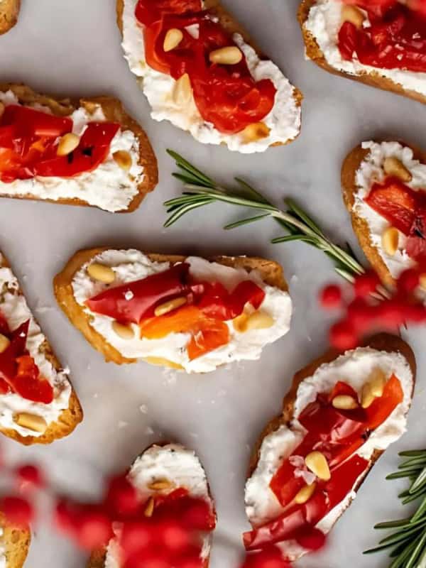 A platter of crostini topped with creamy cheese, roasted red peppers, and pine nuts. Sprigs of fresh rosemary are scattered around for garnish. Bright red berries are blurred in the foreground, adding a festive touch.