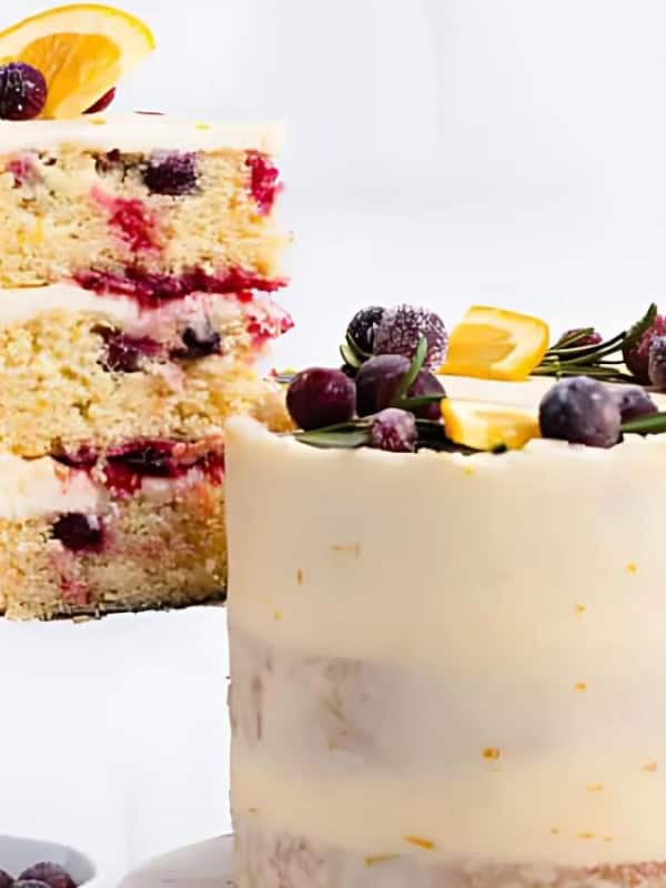 A three-layer orange cranberry cake with white frosting, topped with orange slices, cranberries, and rosemary sprigs.