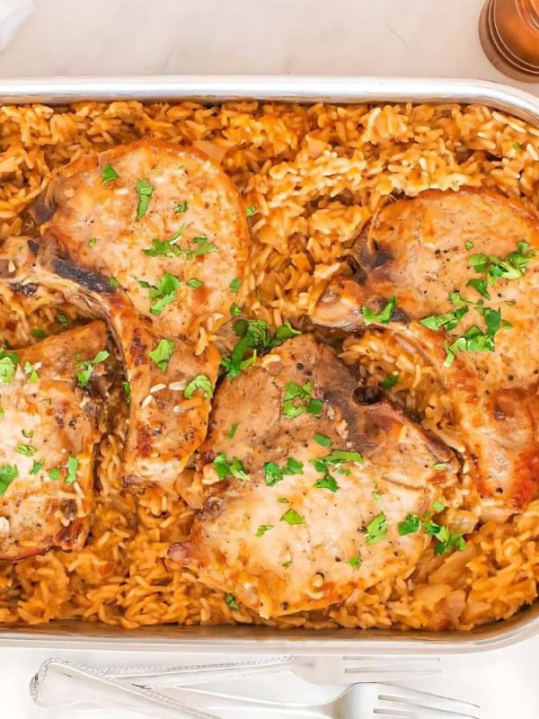 A rectangular baking dish containing four seasoned pork chops on a bed of golden rice, garnished with parsley. Beside the dish are a small bowl of butter, a jar of seasoning, a pepper shaker, and fresh greens.