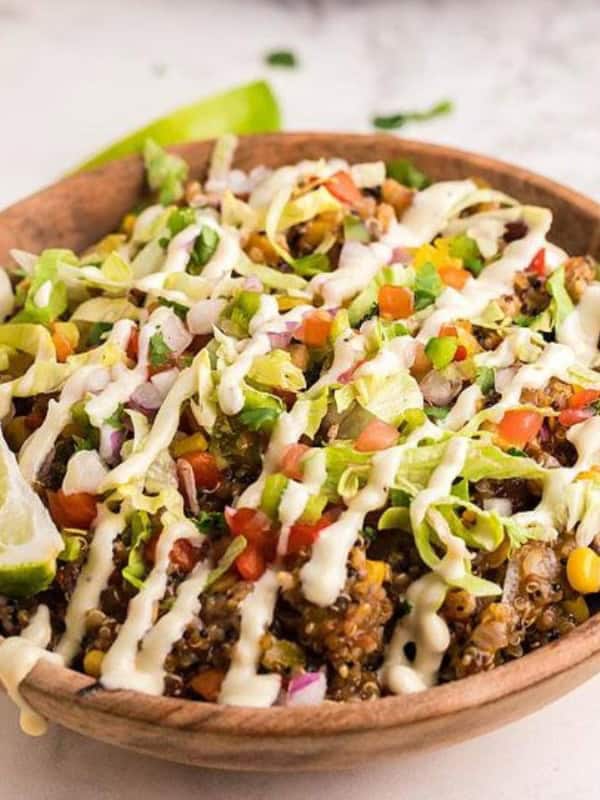 A wooden bowl filled with a vibrant quinoa salad topped with diced tomatoes, onions, corn, and avocado. Drizzled with creamy sauce and garnished with a lime wedge. Additional lime wedges and a small bowl of salsa are in the background.