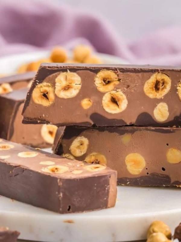 A stack of chocolate bars with hazelnuts embedded, placed on a round marble board. More chocolate bars and a bowl with chocolate spread are in the background, along with a purple cloth. Some scattered hazelnuts are visible.
