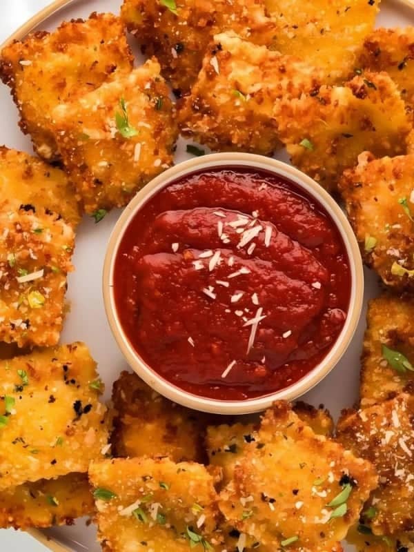 A plate of golden, crispy fried ravioli arranged in a circle, garnished with herbs and grated cheese. A small bowl of marinara sauce is in the center. Fresh parsley and a fork are nearby on a white background.