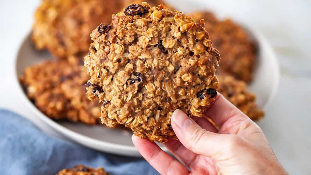 A hand holding an oatmeal cookie with raisins above a plate full of similar cookies. The background includes a blue cloth and blurred items, emphasizing the cookie's texture and ingredients.