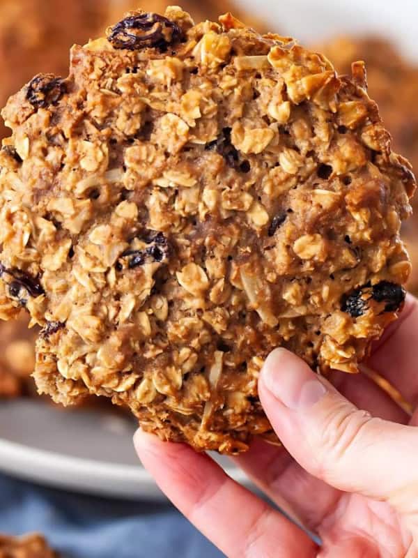 A hand holding an oatmeal cookie with raisins above a plate full of similar cookies. The background includes a blue cloth and blurred items, emphasizing the cookie's texture and ingredients.