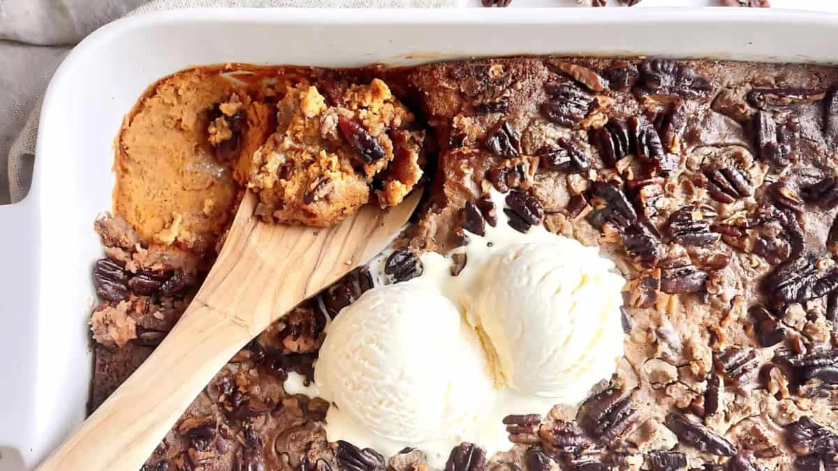 Baked dessert in a rectangular pan with pecans and ice cream scoops on top. A wooden spatula rests in the pan, revealing a portion of the dessert has been served.