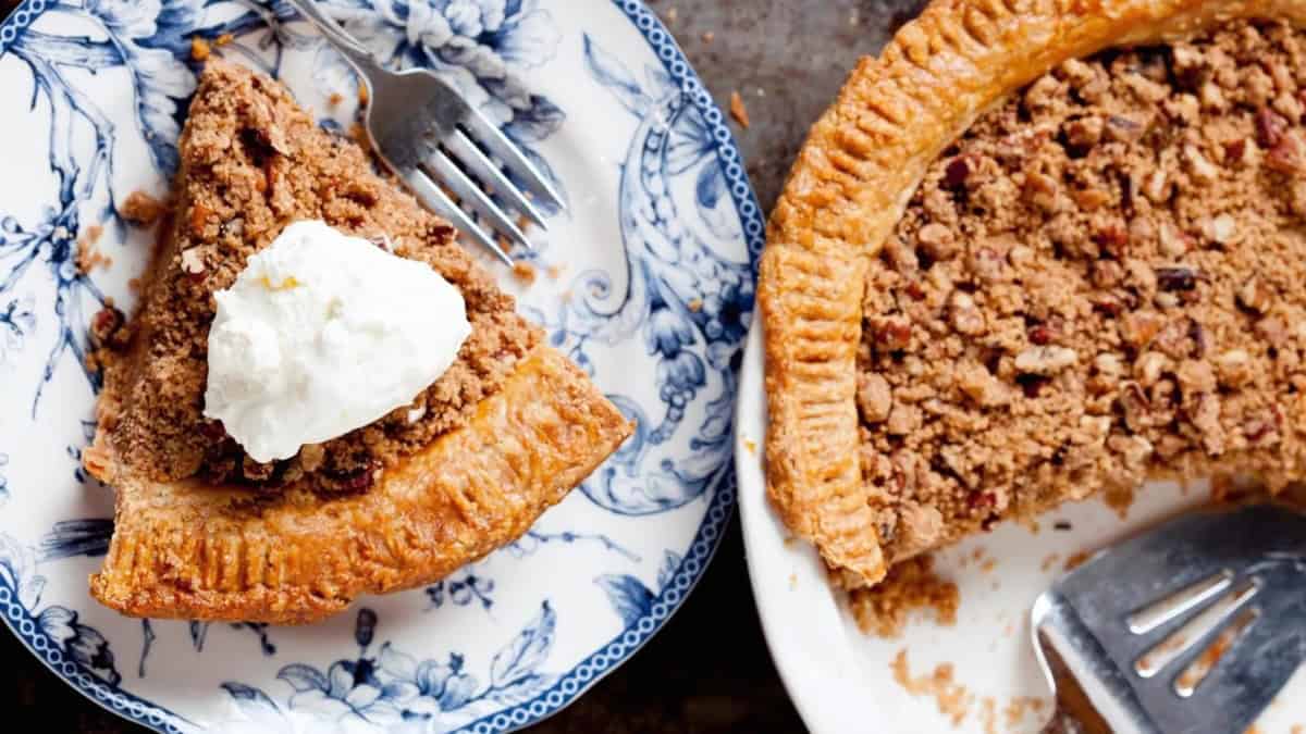 A slice of pecan pie topped with whipped cream on a floral blue and white plate, next to the remaining pie with a pie server on the right. Both sit on a rustic surface, showcasing a golden crust and crumbly pecan filling.