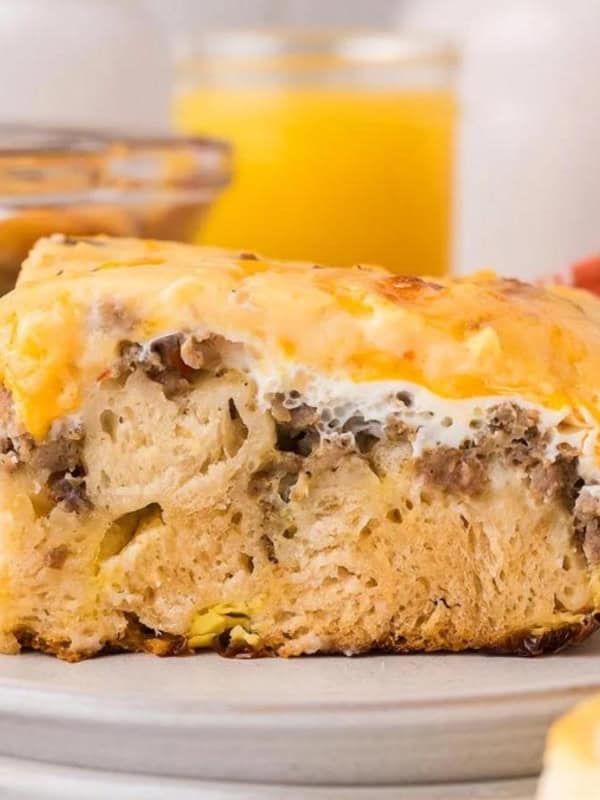 A slice of layered casserole on a gray plate, featuring ingredients like fluffy bread, sausage, and melted cheese. A glass of orange juice and a red-patterned cloth are in the blurred background.