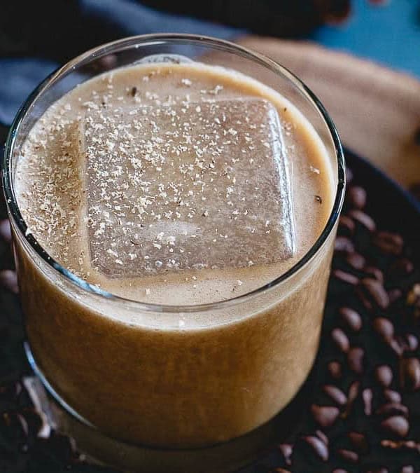 Glass of coconut bourbon nog with a large square ice cube sitting on a plate with coffee beans.