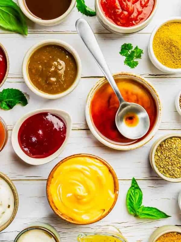 Assorted colorful sauces and spices in small bowls arranged on a white wooden surface. Ingredients include sauces in shades of red, yellow, and brown, surrounded by herbs and various ground spices, creating a vibrant culinary display.