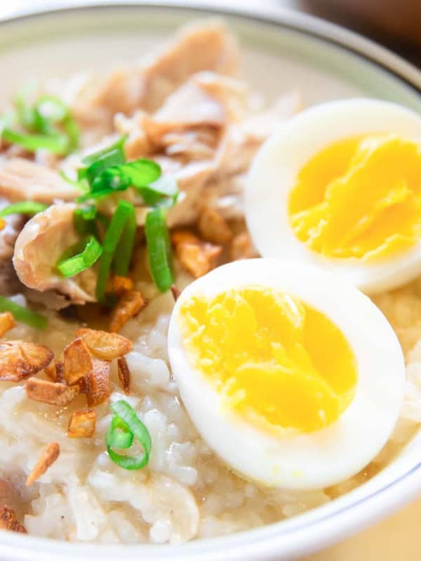 A bowl of rice porridge topped with sliced boiled egg, shredded chicken, fried garlic, and chopped green onions. The eggs are halved, showing bright yellow yolks. The dish is served in a white bowl with a green border.