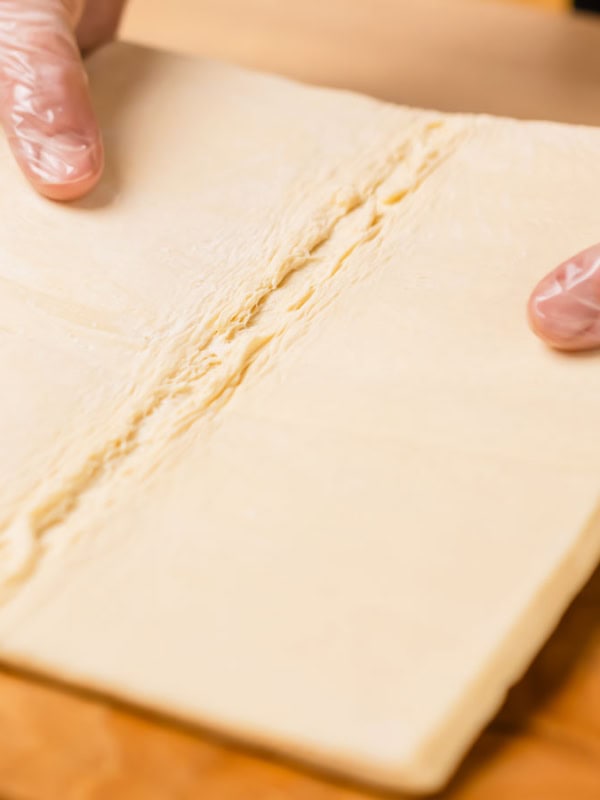 Two gloved hands hold a large square piece of dough on a wooden surface. The dough has a visible fold line down the center. The wooden background contrasts with the smooth, light-colored dough.