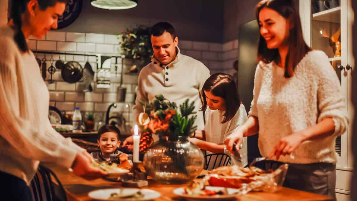 A family of five joyfully gathers around a dining table set with food and a lit candle in a cozy kitchen. They are preparing to share a meal together, wearing warm, casual clothing. The kitchen is warmly lit, with shelves and cooking utensils in the background.