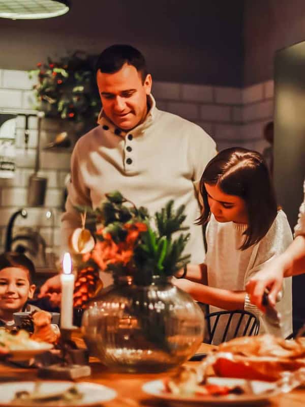 A family of five joyfully gathers around a dining table set with food and a lit candle in a cozy kitchen. They are preparing to share a meal together, wearing warm, casual clothing. The kitchen is warmly lit, with shelves and cooking utensils in the background.