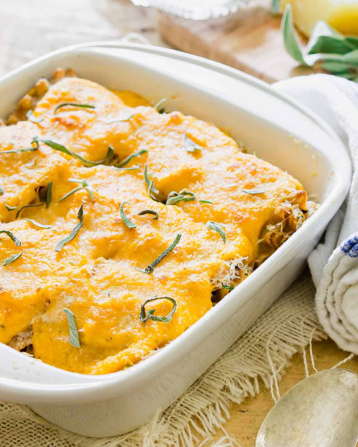 A white rectangular baking dish filled with lasagna rollups topped with a creamy sauce and garnished with sliced fresh herbs. It rests on a rustic cloth next to a folded kitchen towel and spoon.
