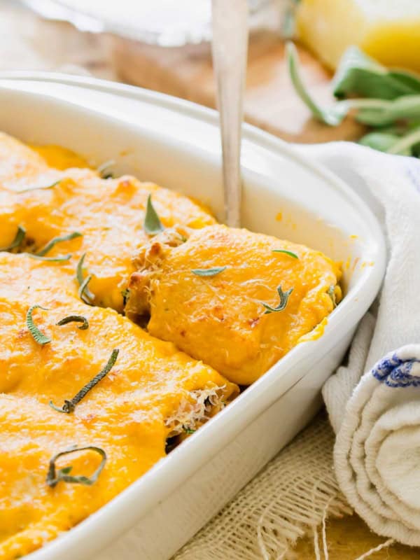A casserole dish filled with lasagna rollups, topped with a creamy butternut squash sauce and fresh sage. A serving spoon is lifting out a portion, showing layers underneath. A white and blue towel is partially visible on the side.