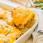 A close-up of a slice of cheesy butternut squash lasagna rollups being lifted from a baking dish. The lasagna is topped with melted cheese and garnished with fresh sage leaves. A white towel is visible in the background.