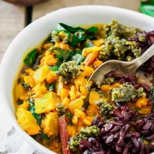 A white bowl filled with lentil stew, featuring vibrant greens, chopped vegetables, topped with dark grains and a pesto-like sauce. A spoon rests in the bowl, with a light-colored fabric underneath.