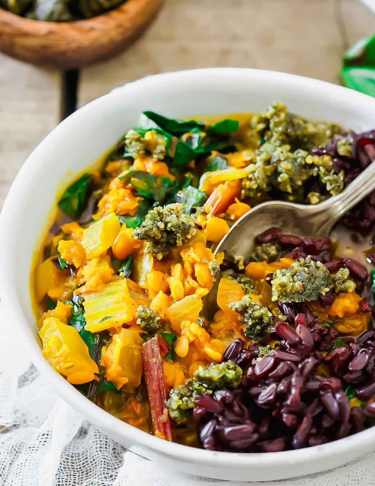 A white bowl filled with lentil stew, featuring vibrant greens, chopped vegetables, topped with dark grains and a pesto-like sauce. A spoon rests in the bowl, with a light-colored fabric underneath.