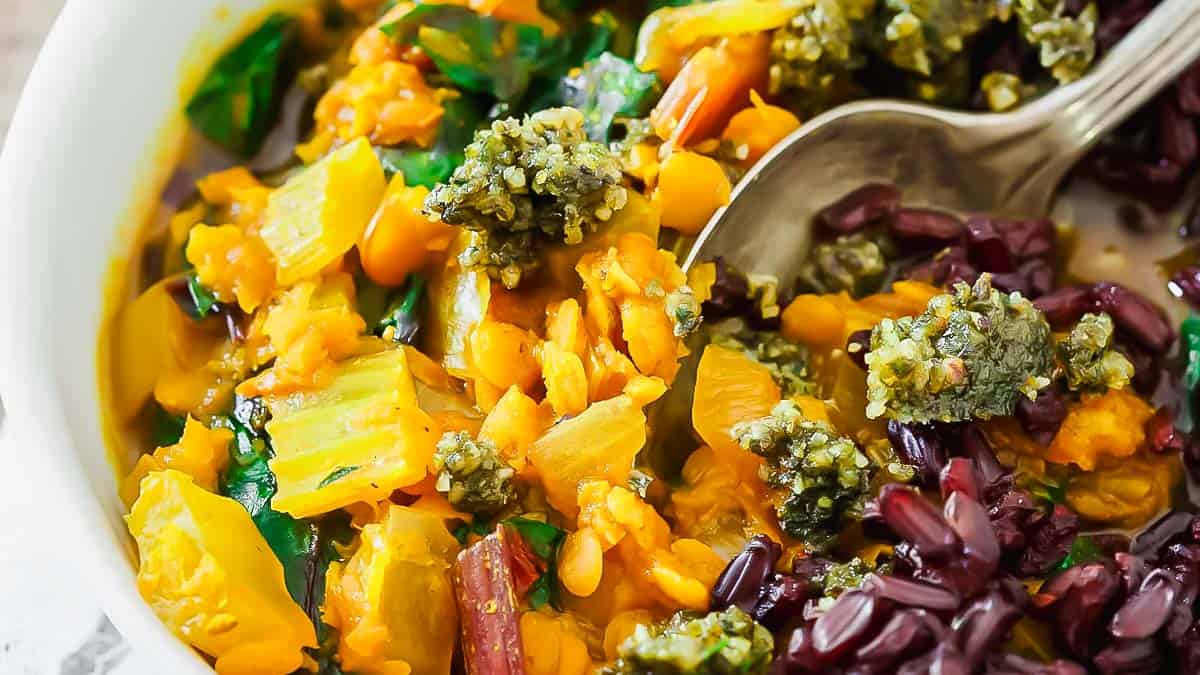 A colorful plate of lentil turmeric stew served with black rice and a pesto sauce. A spoon is placed on the side of the dish. The food appears vibrant and fresh.