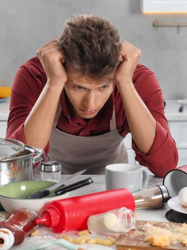 A frustrated person stands in a messy kitchen with ingredients and utensils scattered around. A rolling pin, eggs, and flour are visible on the counter. They appear overwhelmed by the chaos.