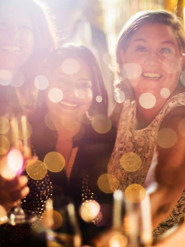 A group of people enjoying a celebration, smiling and holding glasses, with bright, festive bokeh lights in the foreground. The atmosphere is joyful and vibrant.