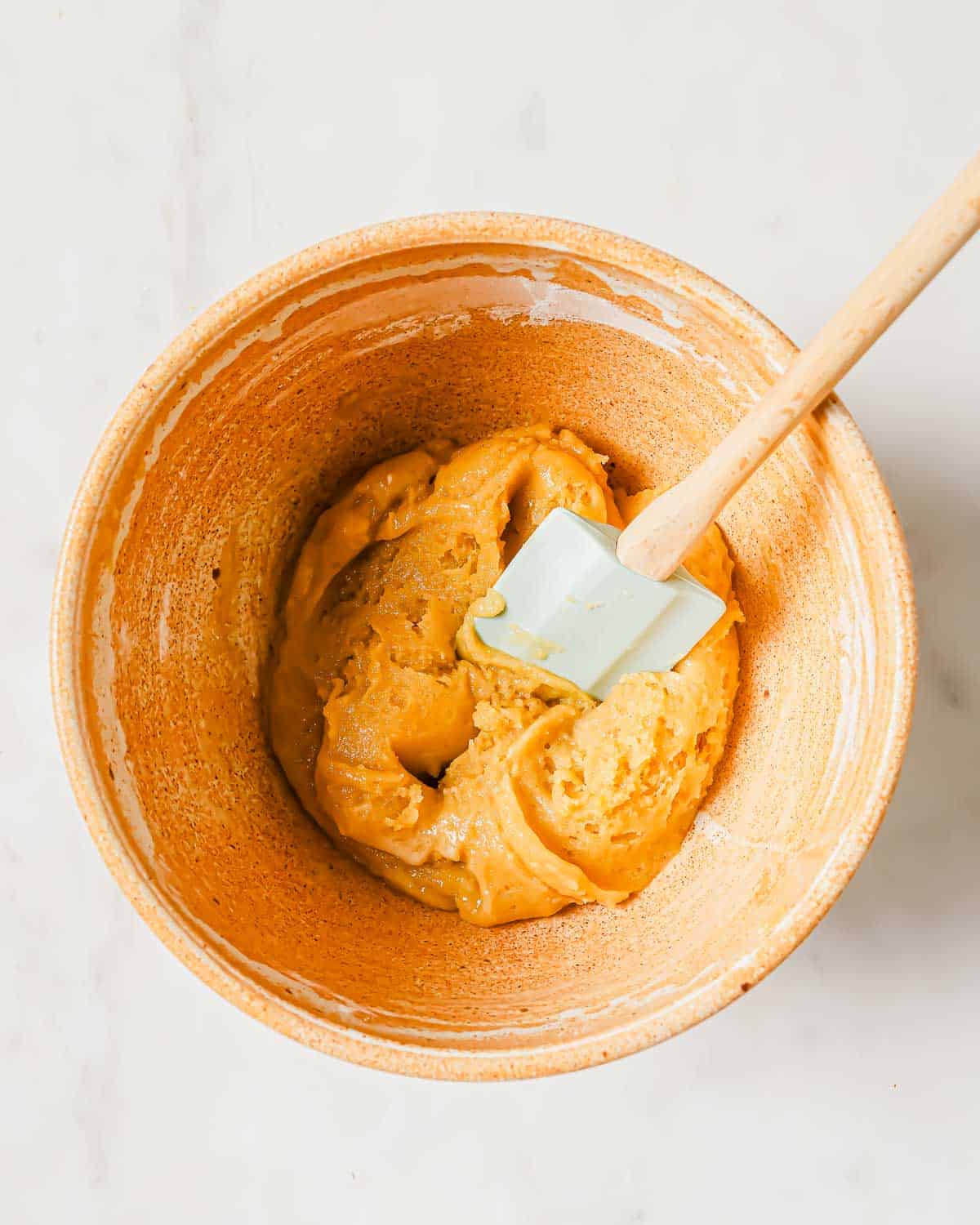 A ceramic bowl with a smooth olive oil cookie batter and a spatula resting inside on a light background.