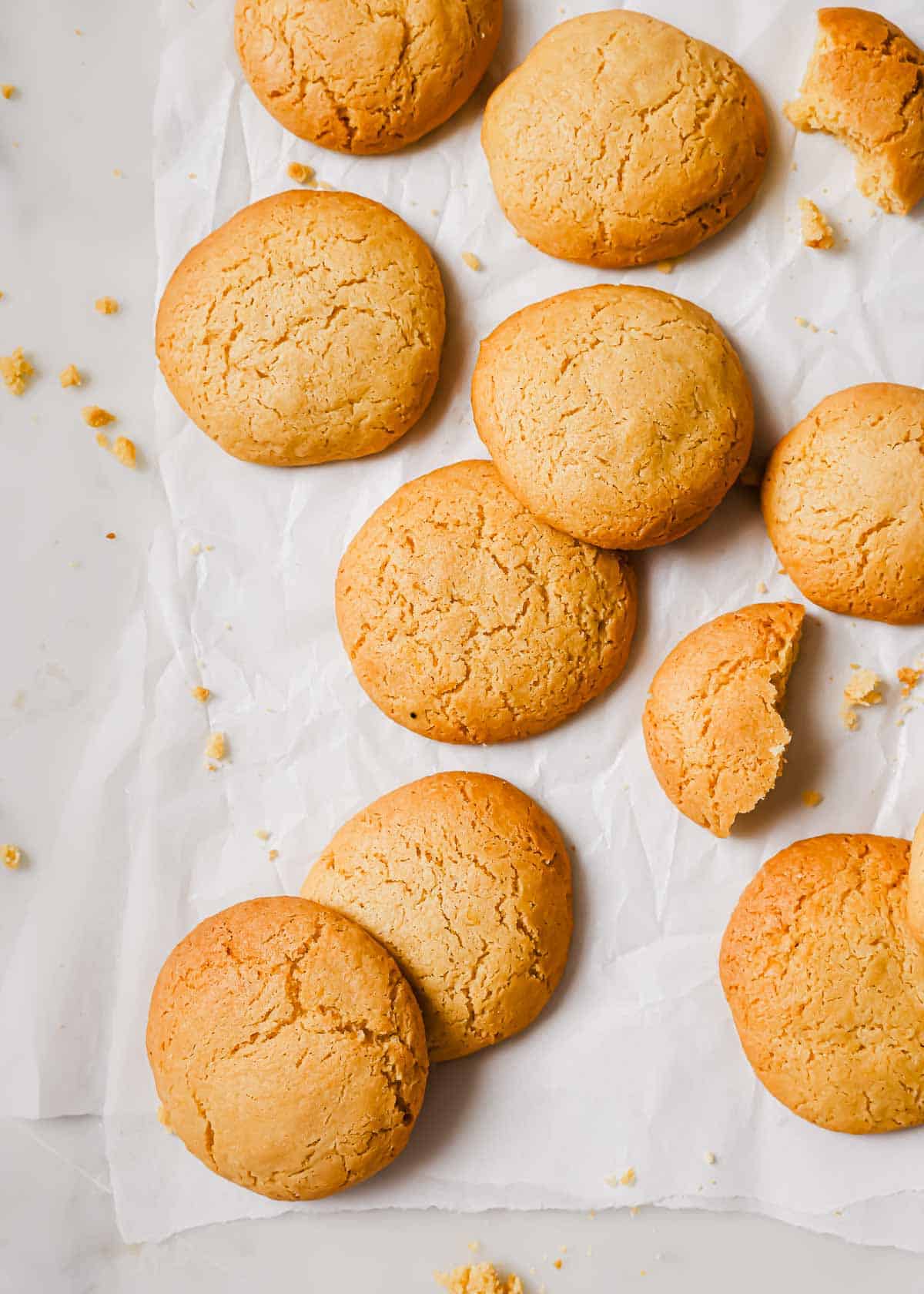 A batch of golden-brown lemon olive oil cookies on crumpled parchment paper. Some cookies are whole, while one is partially broken, revealing a soft interior. Crumbs are scattered on the surface, enhancing the homemade and rustic feel.