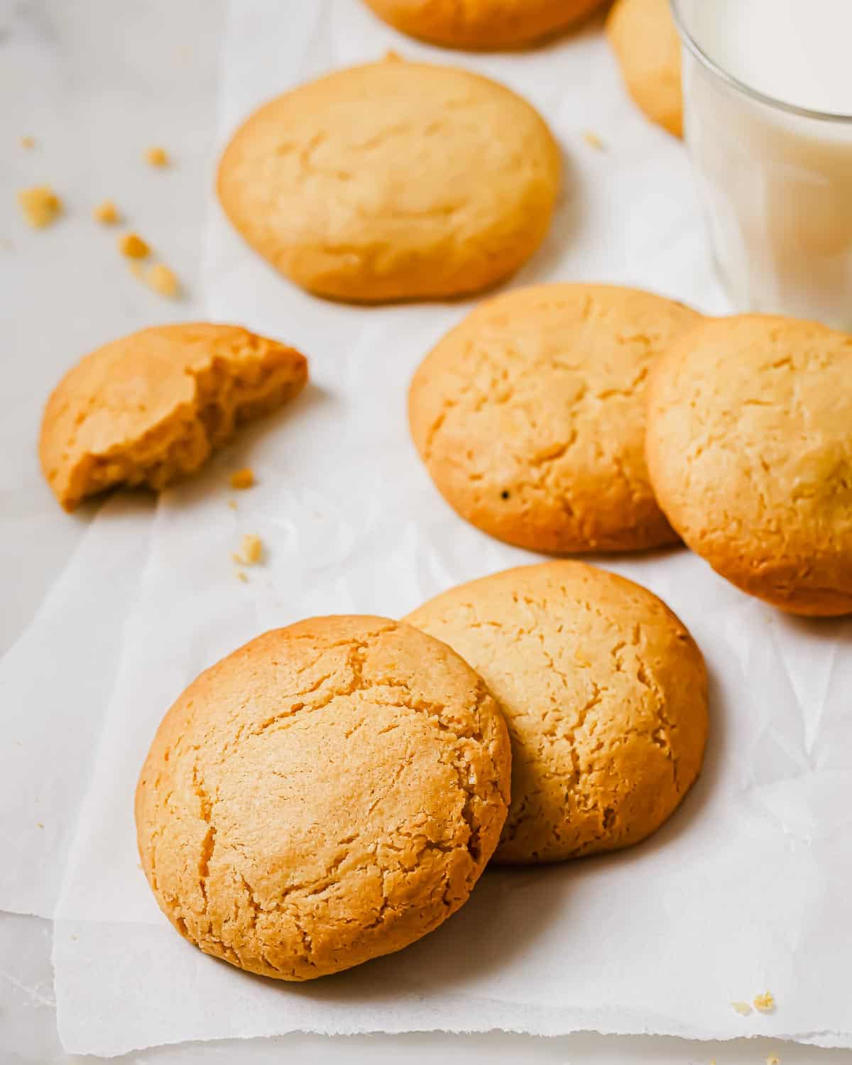Freshly baked cookies on white parchment paper, one with a bite taken out. A glass of milk is placed nearby. Crumbs are scattered around, giving a homely and inviting appearance.