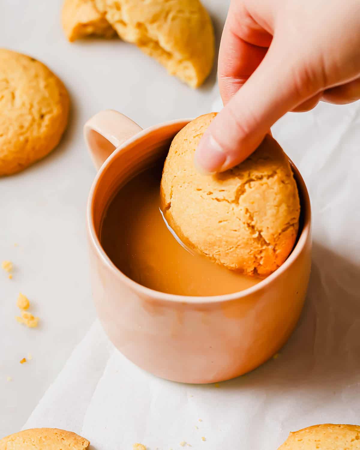 A hand dips a large lemon crinkle olive oil cookie into a pink mug filled with coffee. More cookies are scattered on a light surface around the mug.