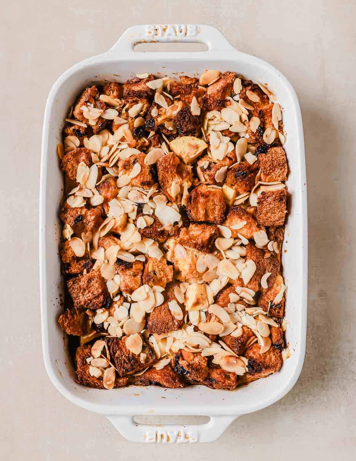 A white casserole dish filled with baked bread pudding topped with sliced almonds and raisins on a light-colored surface.