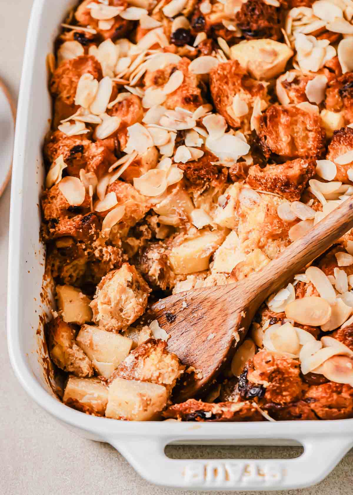 A close-up of a baking dish containing baked bread pudding with apple chunks and sliced almonds on top. A wooden spoon is partially submerged in the dessert, indicating it is being served.