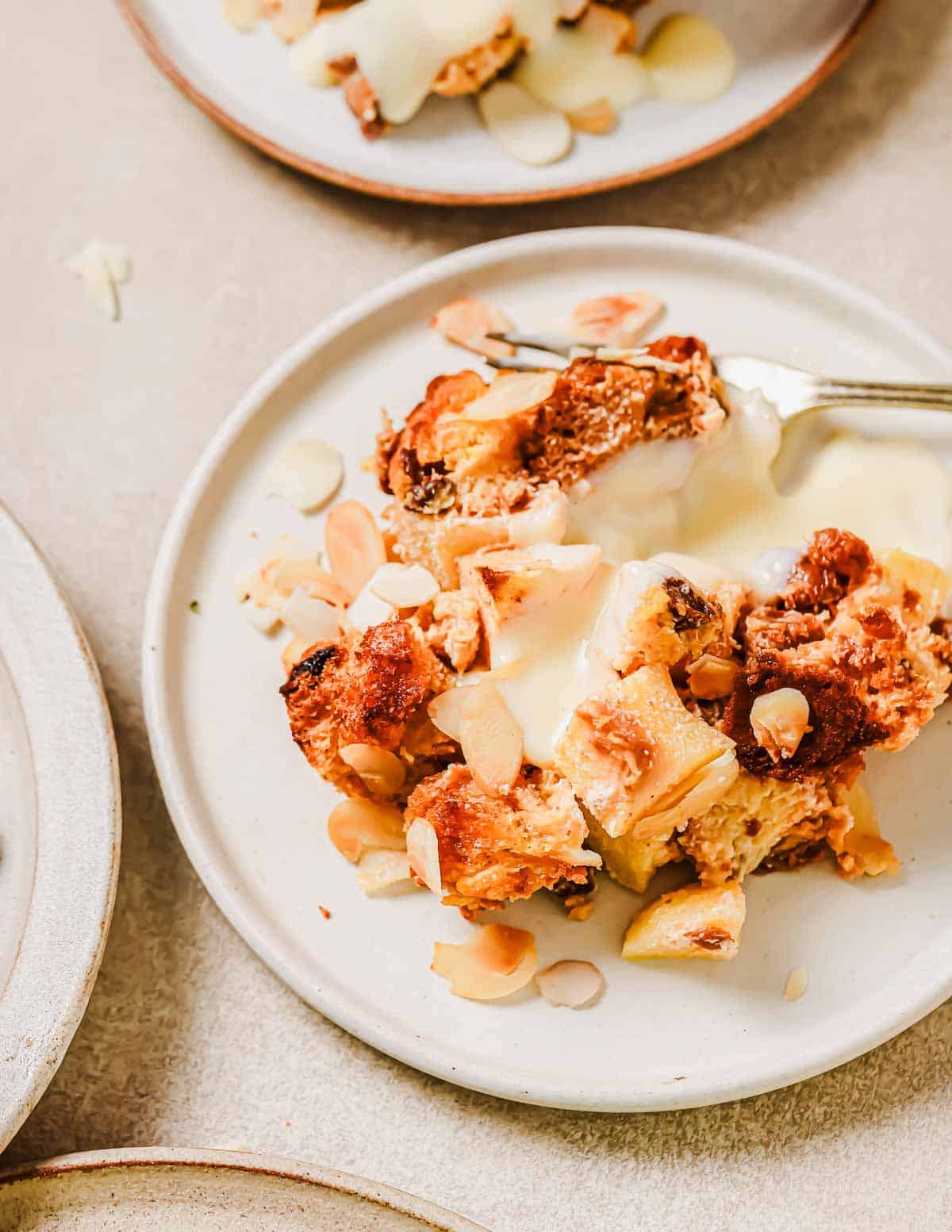 A plate of panettone bread pudding topped with almond flakes and drizzled with cream. A fork rests on the side of the plate, and the dish sits on a light-colored table.