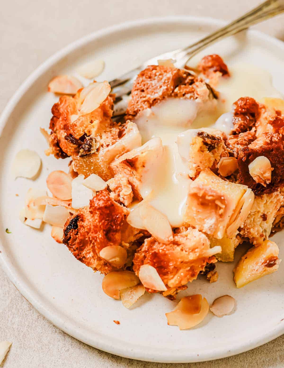 A close-up of a bread pudding dessert on a white plate, featuring a baked mixture topped with custard and sliced almonds. A fork rests on the plate, surrounded by a variety of golden-brown and pale layers.