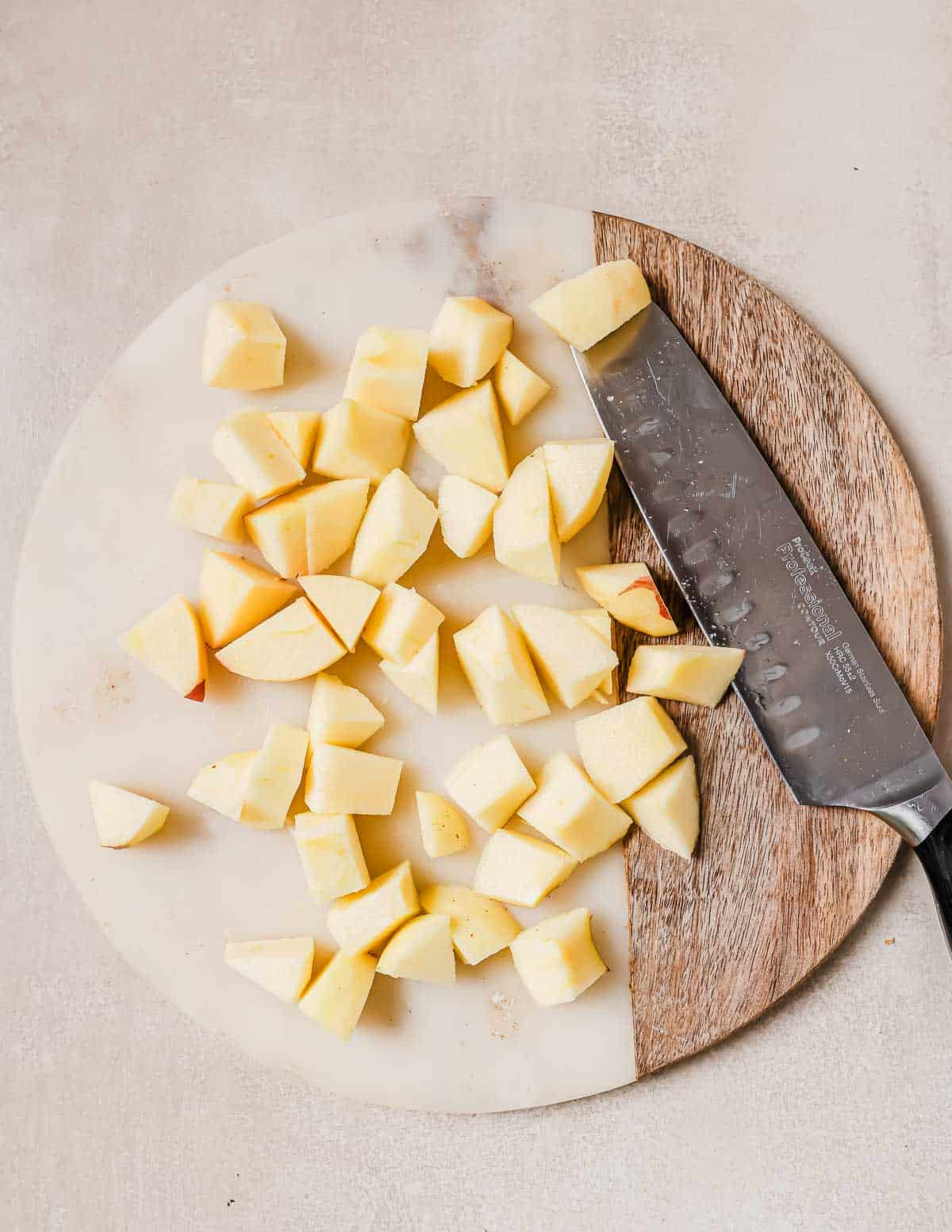 Chopped apple pieces on a round cutting board with a large knife placed beside them. The cutting board has a wood and marble design on a light-colored surface.