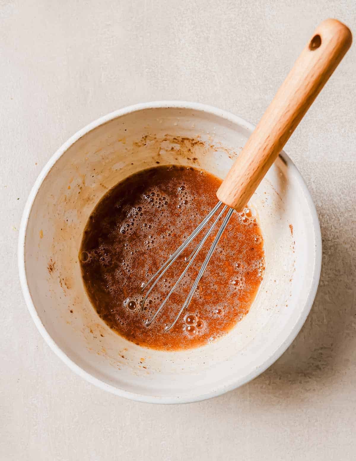 A white mixing bowl containing heated molasses is on a light surface. A whisk with a wooden handle is resting inside the bowl. The mixture appears bubbly.