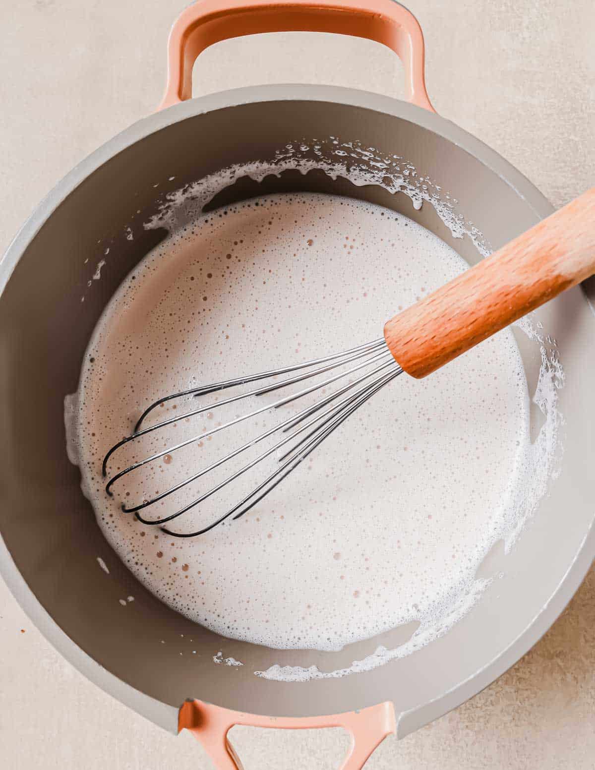Top view of a pot with creamy, frothy milk being whisked. The pot has two handles and is light-colored, with a wooden-handled whisk resting inside. The mixture appears to be smoothly blended.