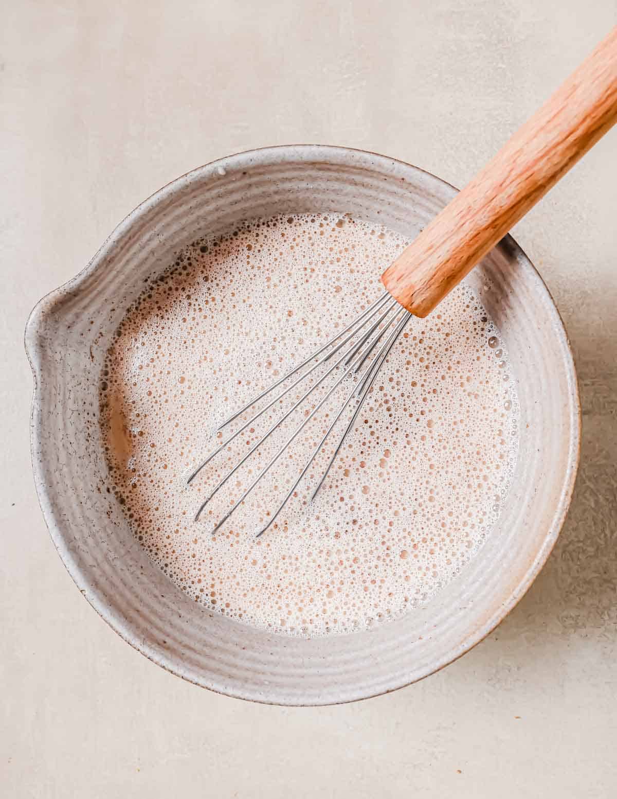 A ceramic bowl with a handle is filled with a frothy milk and spices mixture. A whisk with a wooden handle rests inside the bowl.