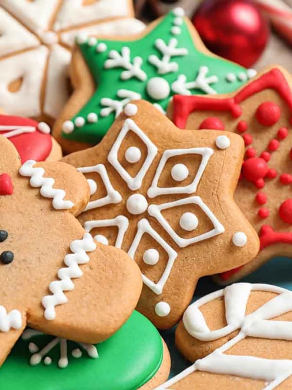 A festive assortment of decorated gingerbread cookies on a blue plate. The cookies include a gingerbread person, snowflakes, stars, and candy canes, with red, white, and green frosting. Candy canes and red ornaments are in the background.