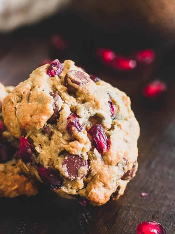 Two cookies studded with chocolate chips and pomegranate seeds are placed on a dark wooden surface. More pomegranate seeds are scattered around, adding a pop of red to the rustic setting. A blurred background with more cookies is visible.