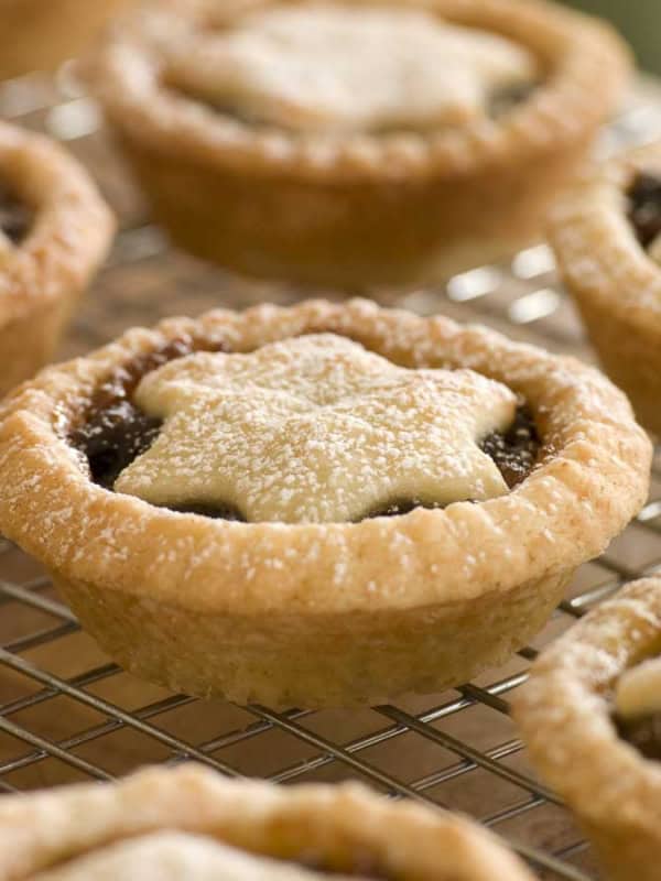 Close-up of freshly baked mince pies on a cooling rack. Each pie is topped with a star-shaped pastry and dusted with powdered sugar. The golden crusts and star toppings give a festive appearance.