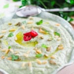 A bowl of eggplant dip garnished with olive oil, sliced red chili, pine nuts, pumpkin seeds, and fresh parsley. A spoon rests beside it, with more fresh parsley in the background.