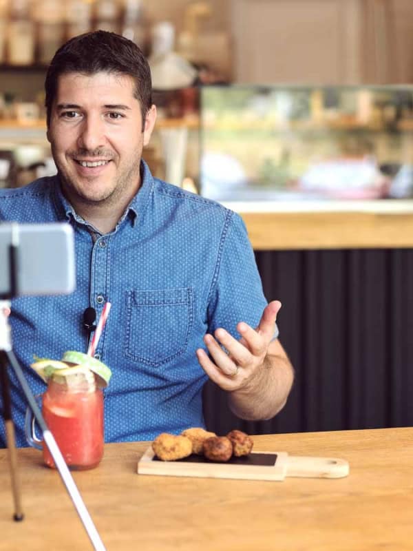 A man in a blue shirt sits at a wooden table in a cafe, recording himself with a smartphone on a tripod. On the table are a mason jar with a red drink and a cutting board with food. A staff member works in the background.