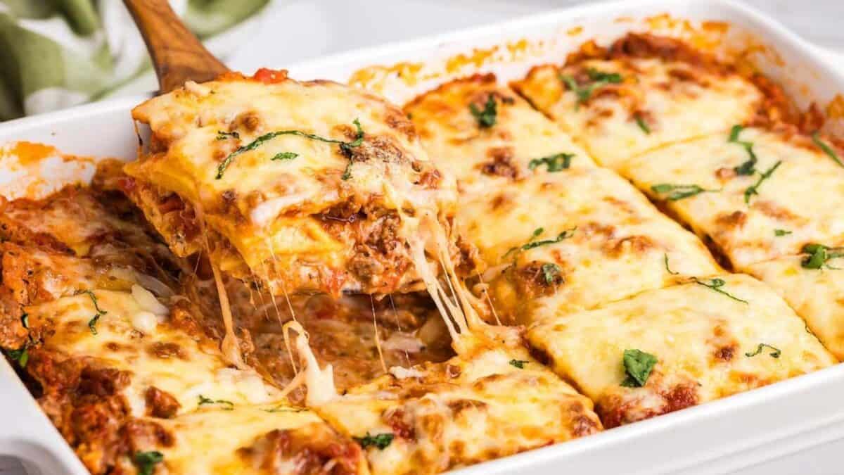 A cheesy lasagna being served from a white baking dish. The slice shows layers of pasta, meat sauce, and melted cheese, with a bit of fresh basil on top. The rest of the lasagna remains in the dish, with gooey cheese stretching.