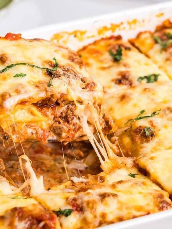 A cheesy lasagna being served from a white baking dish. The slice shows layers of pasta, meat sauce, and melted cheese, with a bit of fresh basil on top. The rest of the lasagna remains in the dish, with gooey cheese stretching.