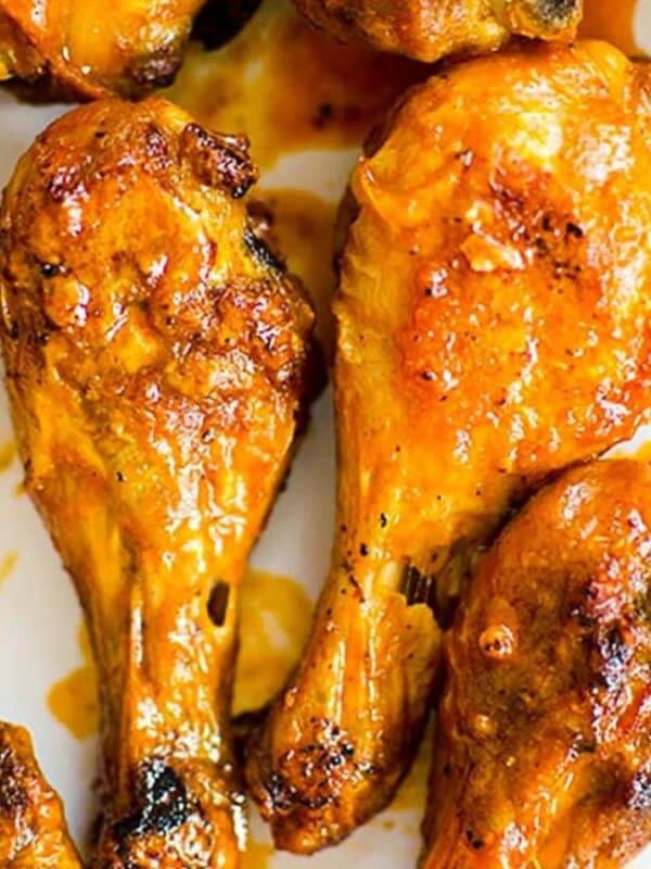 Close-up of golden-brown chicken drumsticks coated in a glossy sauce, arranged on a white plate. The chicken skin appears crispy, and sauce drips slightly, suggesting a flavorful, cooked finish.