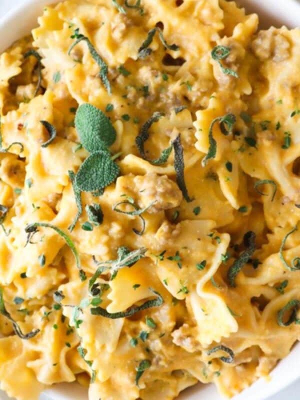 A bowl of creamy bow-tie pasta garnished with chopped herbs. The pasta is mixed with a rich sauce, possibly cheese or cream-based, and is sprinkled with small, fresh green leaves. A woven coaster and fresh herbs are nearby.