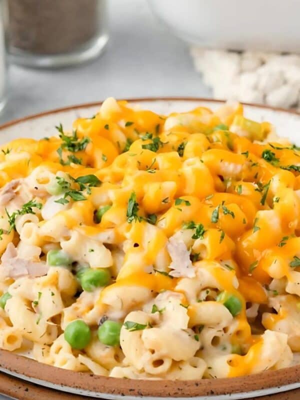A plate of cheesy macaroni casserole with chunks of chicken and green peas, garnished with herbs. A striped napkin, a fork, and a glass container are in the background on a light-colored table.