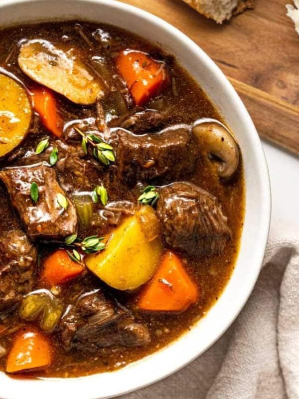 Bowl of beef stew. The stew is dark brown and contains chunks of beef, potatoes, carrots, and mushrooms in a rich gravy. A sprig of thyme is on top. The bowl is white and sits on a beige linen napkin. A wooden cutting board with a loaf of bread is in the upper right corner. The background is a light gray surface.